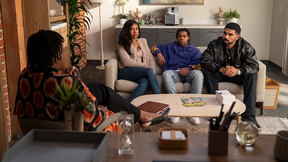 Three people sit on a couch in a modern, warmly lit office, facing another person in a chair, suggesting a therapy or counseling session. The room has plants, shelves, and a coffee table with magazines.