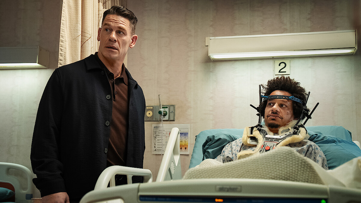 A man stands beside a hospital bed where another man, wearing a neck brace and headgear, lies injured in a medical room with monitors and equipment.
