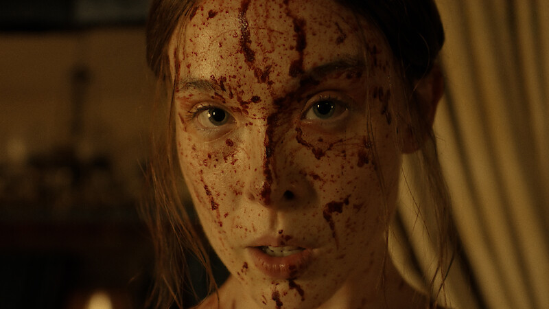 A woman with long brown hair stands indoors with blood splattered across her face, looking intensely forward; the background is dim and softly lit with curtains and indistinct objects.