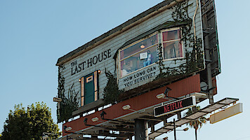 A creative billboard designed as a rundown house with vines, a door, windows, and a sign reading "The Last House" and "How long can you survive?", promoting a Netflix show, set outdoors against a clear blue sky.