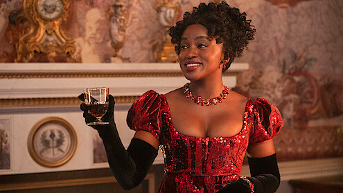 Woman in a red velvet dress and black gloves holds a drink, smiling in an ornate room with a decorated mantle and patterned wallpaper, suggesting a historical or period setting.