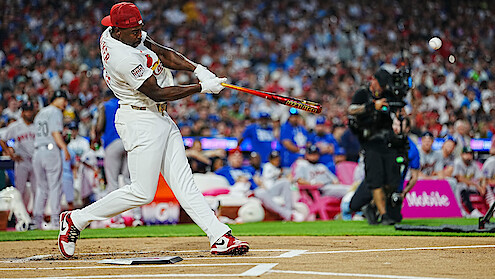 Baseball player in white uniform swinging bat during a game, crowded stadium background with spectators and photographers, action shot captured at home plate.