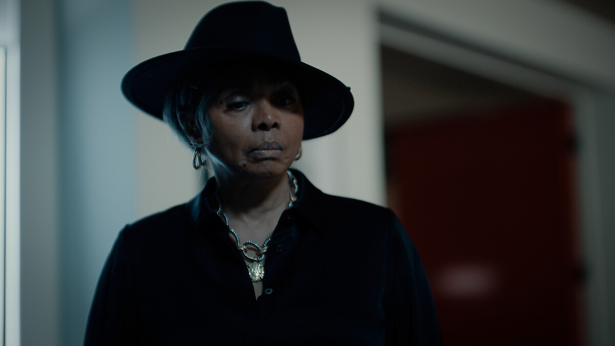 Person wearing a dark wide-brimmed hat and black outfit stands indoors in dim lighting, with a serious expression, silver jewelry visible, and blurred background showing a doorway and red wall.