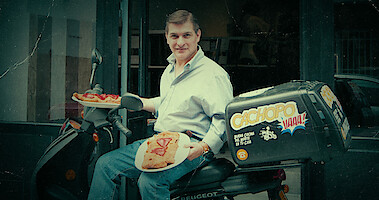 César Román holds plates of food while sitting on a parked motorcycle in an archival photo from the series 'Cooking Up Murder: Uncovering the Story of César Román' 