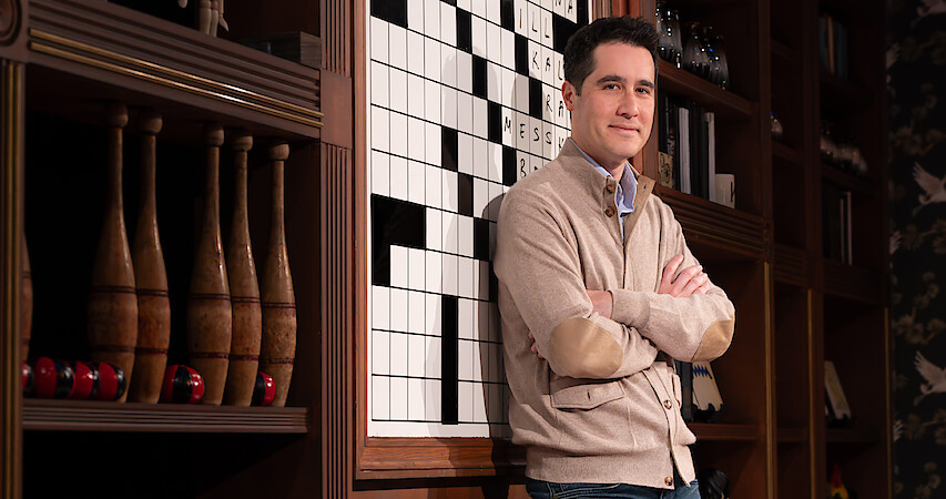 Man in a beige cardigan stands with arms crossed in front of a large crossword puzzle, surrounded by wooden shelves filled with books, decorative bowling pins, and vintage items in a warmly lit library or lounge setting.