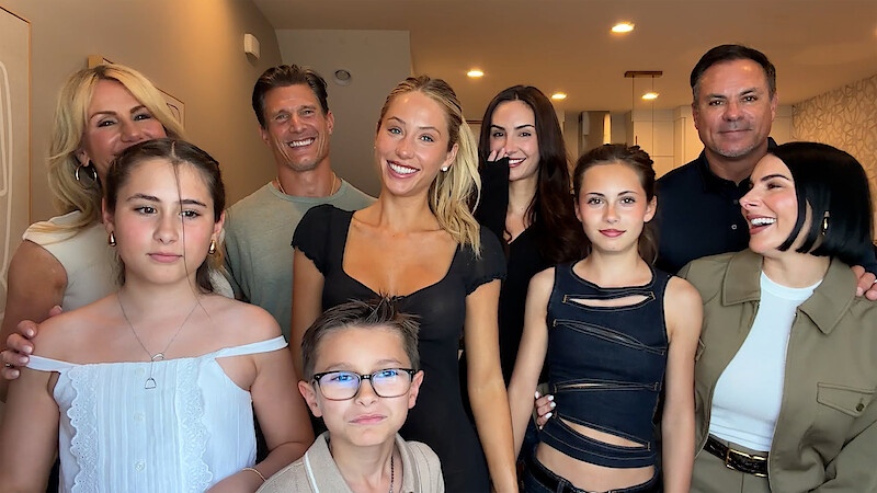A smiling group of adults and children posing together indoors in a well-lit, modern home setting with neutral colored walls and ceiling lights.