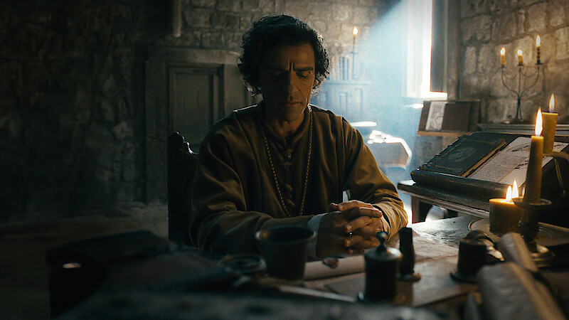 A man sits at a medieval wooden desk in a dimly lit stone room, surrounded by candles, books, and writing tools, with sunlight streaming through a small window, creating a contemplative atmosphere.