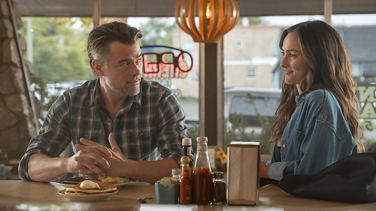  Josh Duhamel as Staten and Minka Kelly as Quinn sit across from each other at a diner table, talking and smiling. Sunlight streams through the windows, and condiments, plates of food, napkins, and a neon sign are visible in a cozy restaurant.