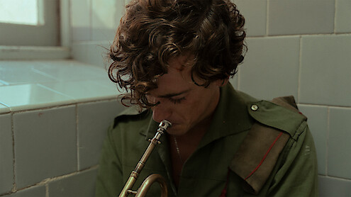 A young man in a green military uniform sits in a tiled room, head bowed as he holds a trumpet to his lips, appearing thoughtful or somber in a softly lit, quiet environment.