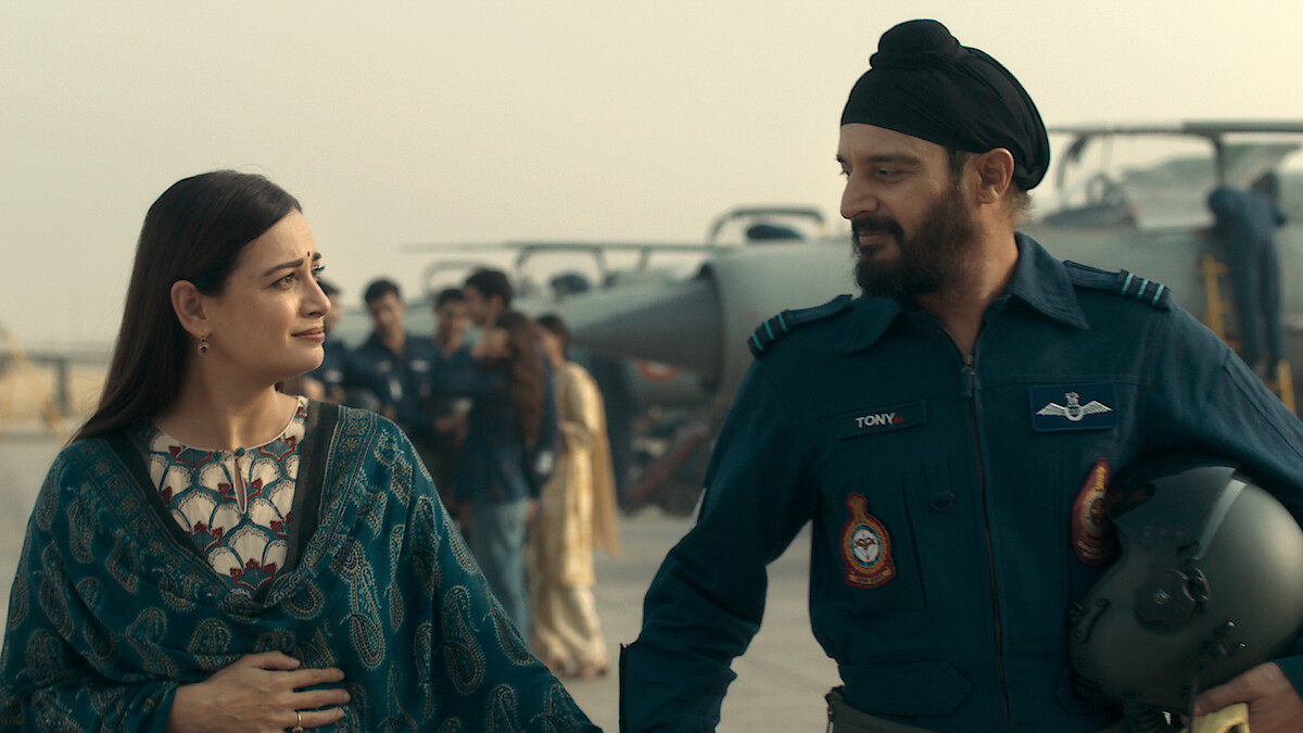A woman and a male Air Force officer in uniform walk together on an airbase, with military aircraft and other personnel visible in the background, evoking a sense of camaraderie and support.