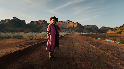 A woman in a pink coat and cowboy hat stands on a path in a Southwestern landscape.