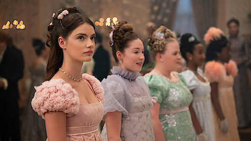 Young women dressed in elegant, pastel-colored Regency-era gowns stand in a row at a formal ball, adorned with floral hair accessories, with candlelight and guests blurred in the ornate, softly lit background.