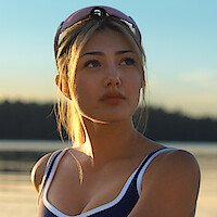 Woman wearing athletic gear and sunglasses looks into the distance outdoors, with a calm lake and forest in the background during sunset.