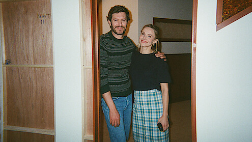 A man and woman smiling, standing close together in a doorway with wooden doors and beige walls, casual clothing, indoor setting with warm lighting.
