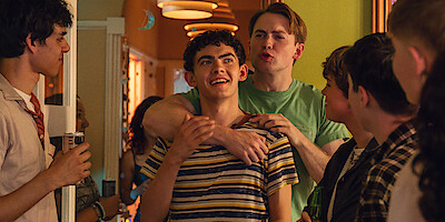 A group of teenagers gather at a lively indoor party. Two boys stand at the center, one smiling while the other playfully wraps an arm around him. The warm, decorated hallway is filled with friends chatting and enjoying themselves.
