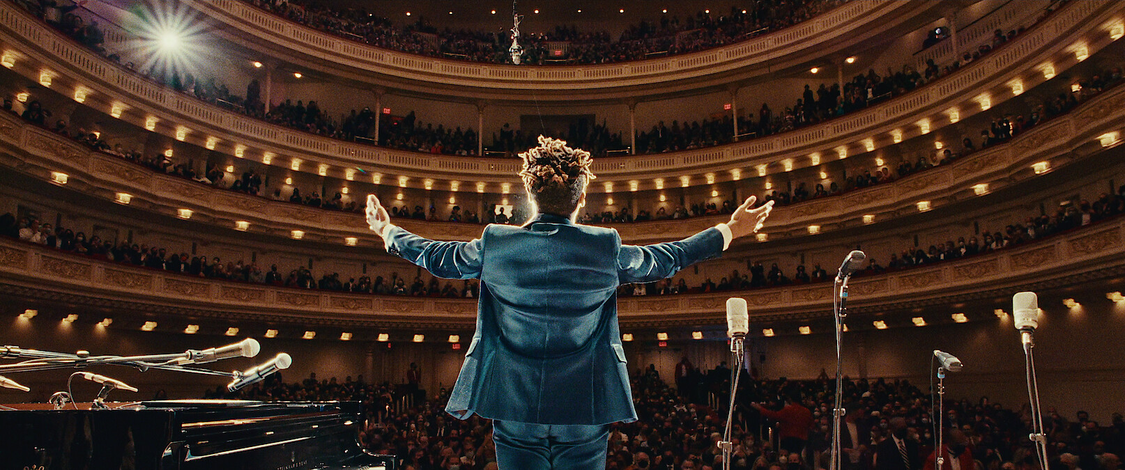 Jon Batiste stands in front of an audience on a stage. 
