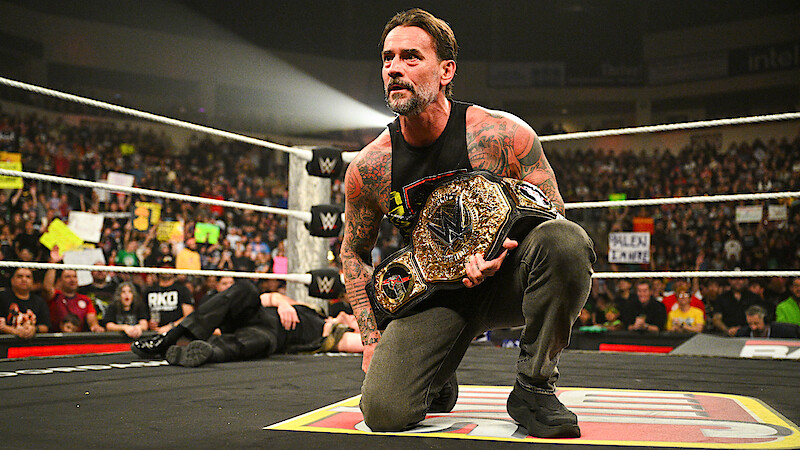 CM Punk kneels in a wrestling ring holding a championship belt, with a cheering crowd and colorful signs in the background during a live event.
