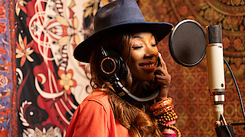 Woman wearing a hat and headphones sings into a microphone in a recording studio with colorful, patterned tapestries in the background.