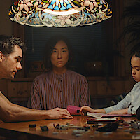 Three people sit quietly around a wooden table under a colorful stained-glass lamp in a dimly lit room; a man to the left, a woman in the center, and a child on the right focused on drawing.
