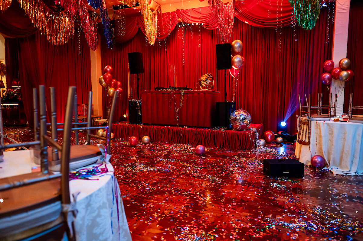Um salão em tons de vermelho onde uma festa acabou de terminar tem papel picado e adereços brilhantes por toda parte