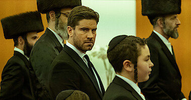 Kevin Janssens, wearing a suit, faces the camera among a group of Orthodox Jewish men wearing shtreimels. 
