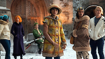 Six people in colorful, winter outfits stand in a snowy courtyard before a large gate, appearing alert or contemplative. The setting is medieval with stone walls and heavy wooden doors.