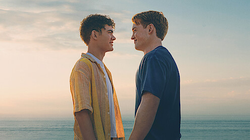Two young men stand facing each other, smiling, at the beach during sunset with the ocean and sky in the background.