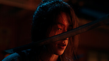 A person with long hair and a cut on their forehead is in a dimly lit, dramatic setting, looking forward with a sword held close to their face, suggesting tension or confrontation.