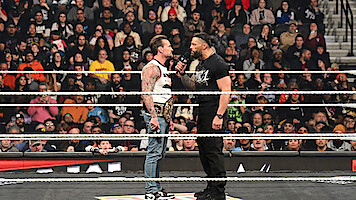 Two wrestlers face off in a packed arena wrestling ring, one holding a championship belt and the other speaking into a microphone as a large audience watches the dramatic confrontation.