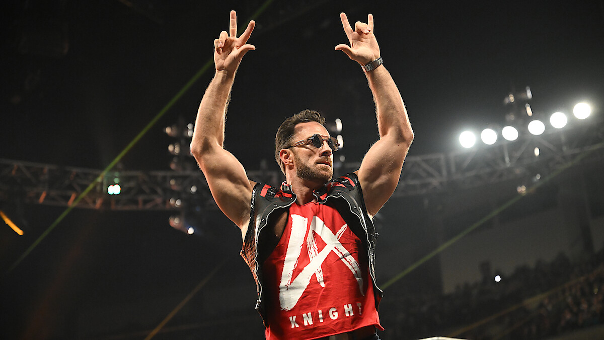 A muscular man wearing sunglasses and a red vest stands in an arena with bright lights, raising both arms and making hand gestures, with an audience and stage lighting visible in the background.