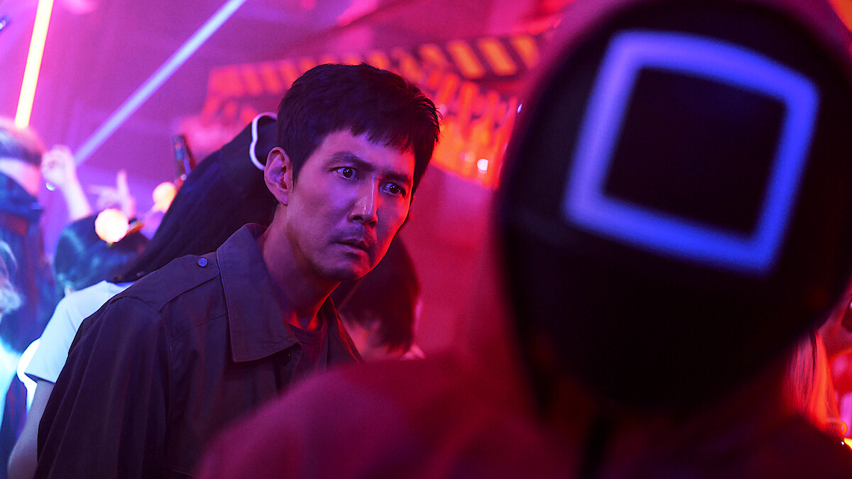 Lee Jung-jae as Seong Gi-hun shows a concerned expression while looking at someone wearing a red jumpsuit and mask in Season 2 of 'Squid Game'