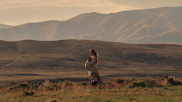 Pregnant woman in a long dress stands in a grassy field with wildflowers, set against a backdrop of rolling hills and mountains during golden hour, creating a serene and natural outdoor scene.
