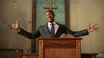 Man in a suit stands at a wooden pulpit with open arms, smiling in a church with a Bible and cross in the background, green and beige walls, and natural lighting.
