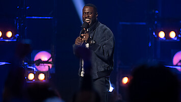 Comedian performing on stage with a microphone, smiling and interacting with an enthusiastic audience in a dark, spotlight-lit comedy club setting.
