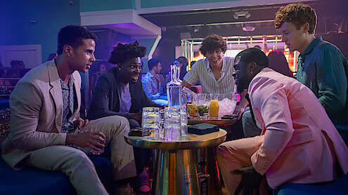 Five men sitting around a table with drinks, laughing and talking in a lively nightclub with colorful lighting and a modern, vibrant atmosphere.