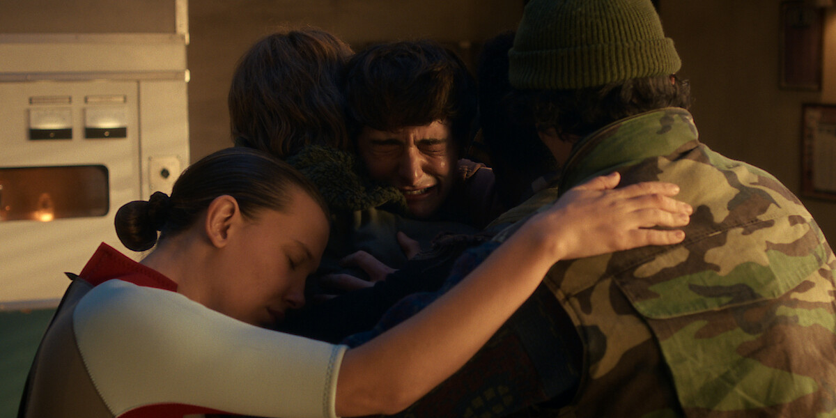 Eleven, Jonathan Byers, Will Byers, and Mike Wheeler sharing an emotional group hug indoors, with eyes closed, in a warmly lit room.