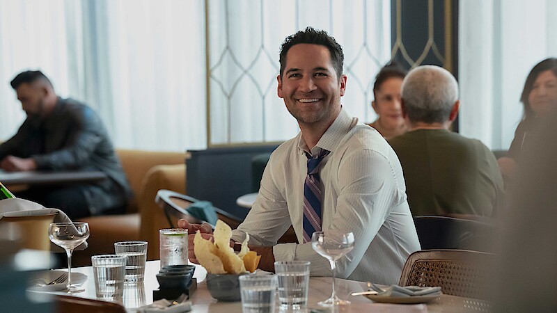 Smiling man in dress shirt and tie sitting at a restaurant table with chips, water glasses, and wine glasses, with other people dining in the background. Bright, modern interior with natural light.
