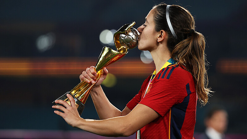 Soccer player in red jersey kisses gold trophy on stadium field, celebrating victory under stadium lights, wearing medal, hair tied back, background out of focus.