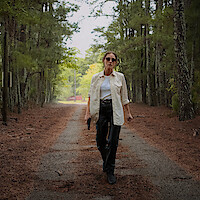 Woman in sunglasses and casual clothes walking down a forest path holding a handgun, surrounded by tall pine trees, with a red structure visible in the background.