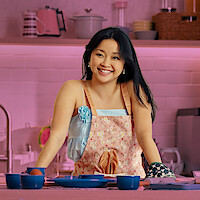 Lara Jean, played by Lana Condor, smiles in a pastel pink kitchen, wearing a floral apron and surrounded by cozy decor.
