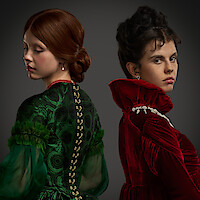 Two women in elaborate period costumes, one in green and one in red, stand back to back against a dark background. Their serious expressions and elegant dresses create a dramatic, tense, and formal mood.