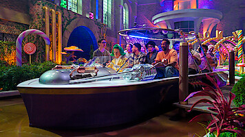 A group of people ride together in a boat through a colorful, whimsical indoor setting with candy decorations, glowing lights, lush plants, and fantasy architecture.