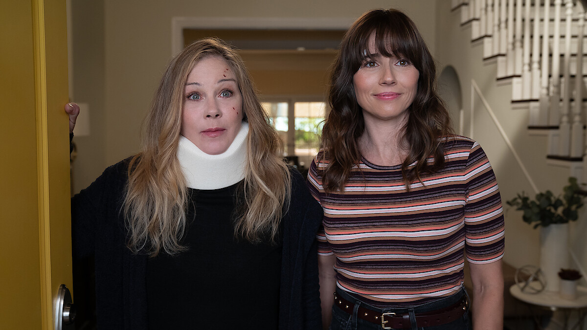 Two women standing in the entryway of a house; one wears a neck brace and dark sweater, the other wears a striped shirt. They stand near an open yellow door, with a staircase and potted plant visible in the background.