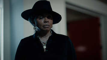 Person wearing a dark wide-brimmed hat and black outfit stands indoors in dim lighting, with a serious expression, silver jewelry visible, and blurred background showing a doorway and red wall.
