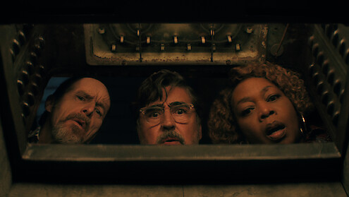 Three adults, two men and one woman, peer with curiosity and surprise into an open oven, as seen from inside the oven looking out in a dimly lit kitchen setting in 'The Boroughs.'