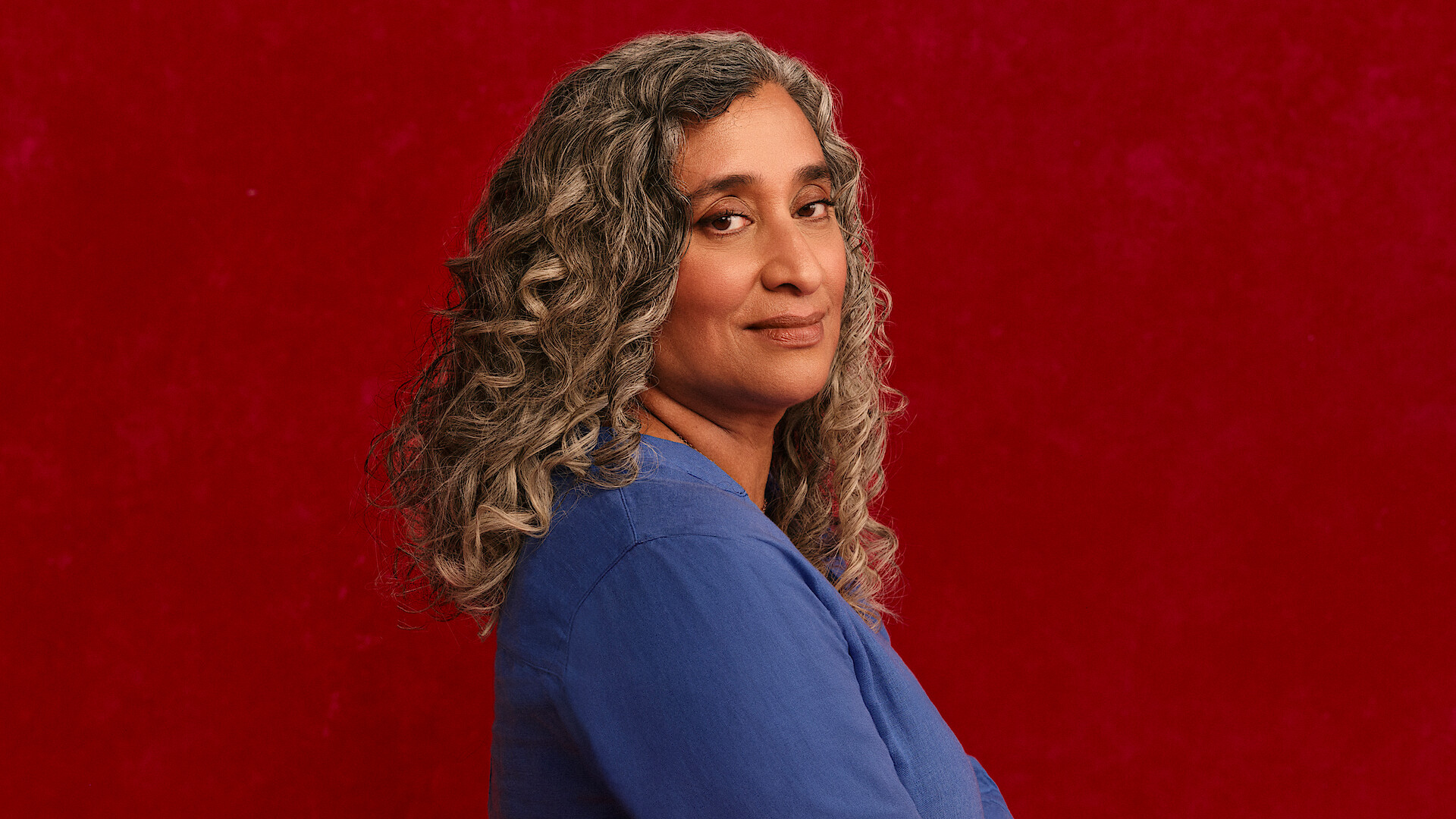 Geeta Gandbhir, director of The Perfect Neighbor, stands in front of a rich red backdrop with beautifully set curls in her graying hair, head turned towards the camera with a confident expression on her face.