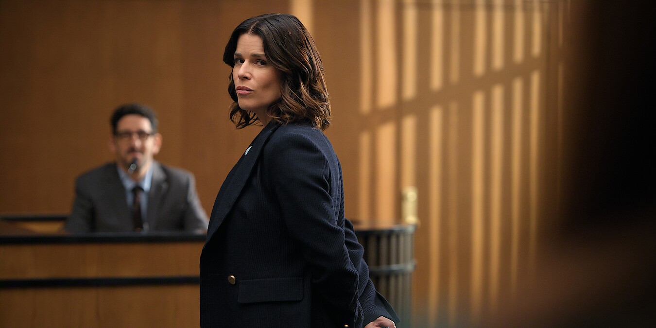 A woman in a dark blazer stands in a courtroom, looking serious, with a man seated at a witness stand in the background, wood panel walls and dramatic light through windows.