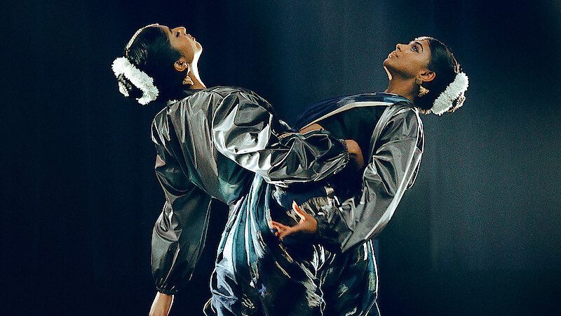 Two dancers in matching shiny costumes perform on stage with dramatic lighting, facing away from each other and leaning back-to-back, wearing white floral hair accessories in a dark, theatrical setting.
