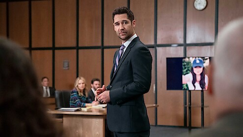 Man in suit stands in courtroom, addressing others. People sit at a table in the background. A digital screen shows a woman’s photo. Wood panel walls and a clock indicate a formal court setting.