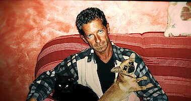A with an angry expression man sits on a couch with a small dog in 'The Yara Gambirasio Case: Beyond Reasonable Doubt'